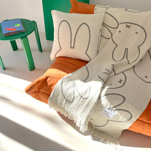 Cream Miffy cotton blanket with fringed edges and gray print, draped over an orange sofa.