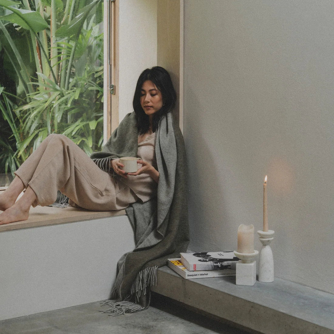 A woman sits on a window ledge in a minimalist room, wrapped in a gray shawl and wearing beige pants and a striped top, holding a cup. A lit candle, books, and a small vase sit on the windowsill next to her, with green plants visible outside the window.