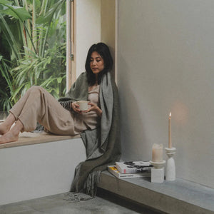 A woman sits on a window ledge in a minimalist room, wrapped in a gray shawl and wearing beige pants and a striped top, holding a cup. A lit candle, books, and a small vase sit on the windowsill next to her, with green plants visible outside the window.