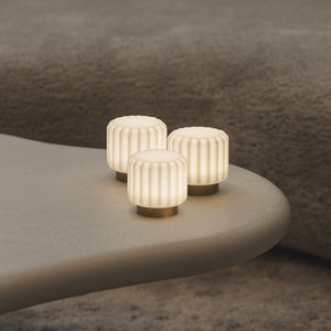 Three small ribbed cylindrical table lamps with warm glowing white shades and gold bases, arranged on a curved light beige side table in a modern living room setting.