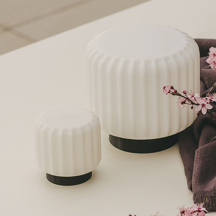 A styled tabletop scene with a large glass jar filled with dark red liquid and botanicals, two small ribbed white containers, a bowl of dark preserves or fruit, and two glasses of red drink, arranged on a white table with a dark cloth and pink blossoms.